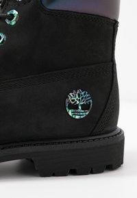 Schwarzer Wildlederstiefel mit einer strukturierten Oberfläche und einer dicken Gummisohle. Besitzt ein holografisches Logo an der Seite, das die intricaten Designdetails hervorhebt.