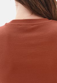 Primer plano del cuello y la parte superior de la espalda de una persona que lleva un suéter de punto acanalado en color óxido y tiene el cabello largo y castaño.