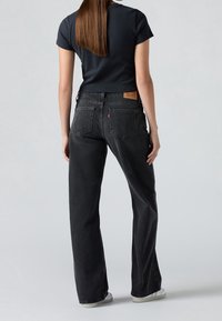 Femme aux longs cheveux bruns portant un t-shirt court noir, un jean taille haute foncé et des baskets blanches, debout dos à la caméra.