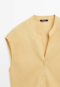 Camicia in lino a maniche corte e scollo a V in un colore beige chiaro, con una texture liscia e due bottoni in legno sul davanti.