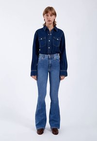 Blå denimskjorte med to brystlommer og knappelukning foran, kombineret med højtsiddende, vide blå jeans og brune støvler.