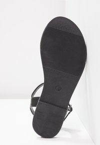 Semelle de sandale noire avec motif de grip texturé, bride de cheville visible, sur fond blanc.