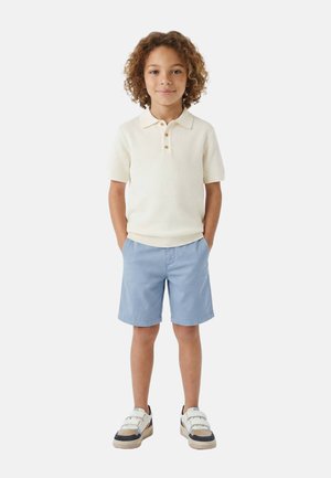Enfant aux cheveux bouclés portant un polo crème à manches courtes, un short bleu clair et des baskets blanches, debout, les mains dans les poches.