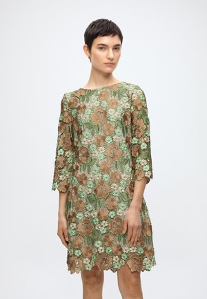 Γυναίκα που φοράει διαφανές φόρεμα μέχρι το γόνατο με καστανή και πράσινη κεντημένη λουλουδένια διακόσμηση και μανίκια τριών τετάρτων, στέκεται μπροστά σε λευκό φόντο.