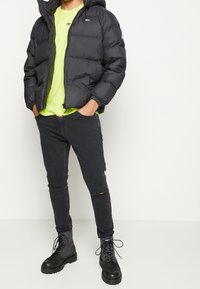 Sort pufferjakke med hætte og lynlås, parret med en neongrøn t-shirt, sort slidte jeans og sorte snørebåndsstøvler.