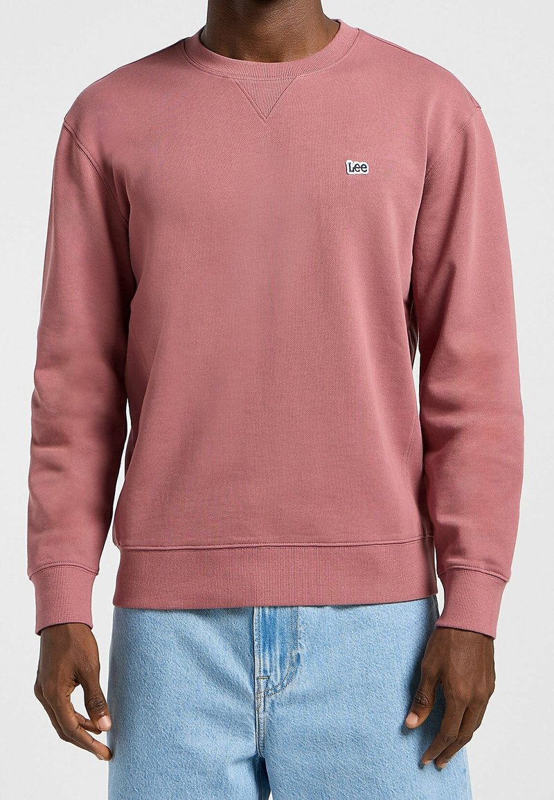 Homme portant un sweat-shirt Lee couleur rose poussiéreux avec des poignets et une ceinture côtelés, associé à un jean en denim bleu clair.