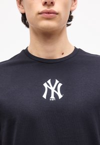 Κοντινό πλάνο ενός ατόμου που φοράει μαύρο πουκάμισο με ένα λευκό στυλιζαρισμένο λογότυπο "NY" στο κέντρο του στήθους.