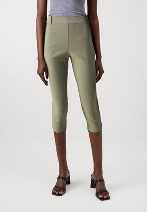 Modèle féminin portant un pantalon court ajusté vert clair, un haut gris sans manches et des sandales noires à talon bloc ouvert, debout sur fond blanc.