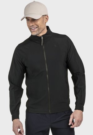 Mann trägt eine beige Baseballkappe und eine schwarze Reißverschlussjacke, lächelt und hat die Hände entspannt an den Seiten, vor einem schlichten Hintergrund.