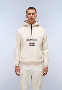 Felpa beige con zip frontale, caratterizzata da un logo e una toppa a forma di bandiera. Tessuto morbido, vestibilità comoda, abbinata a pantaloni cargo beige chiaro.