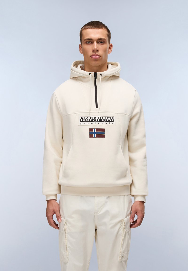 Felpa beige con zip frontale, caratterizzata da un logo e una toppa a forma di bandiera. Tessuto morbido, vestibilità comoda, abbinata a pantaloni cargo beige chiaro.