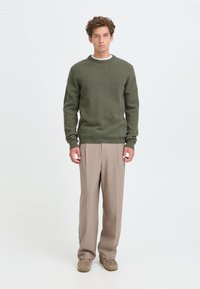 Junger Mann mit lockigem Haar, der einen olivgrünen Pullover, beige Hose und braune Schuhe trägt, steht vor einem einfarbigen hellen Hintergrund.