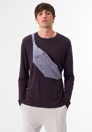 Lila Langarmshirt mit einer grauen Umhängetasche in kurviger Form, glatter Textur, zwei blauen Punkten und einer metallischen Schlaufe.