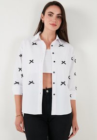 White button-up shirt with black embroidered bows, three-quarter sleeves, and a smooth texture, paired with a cropped top underneath.