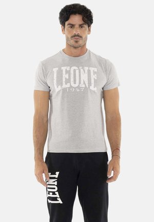 Man met donker haar die een grijs T-shirt draagt met "LEONE 1947" en een zwarte broek met "LEONE" bedrukt op het linkerbeen, staande tegen een witte achtergrond.