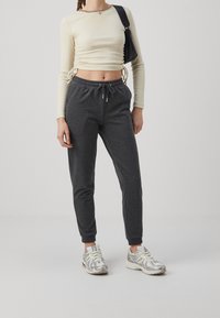 Kvinna iförd en beige långärmad kort topp, mörkgrå joggingsbyxor, silvervita sneakers och bärande en svart axelväska.