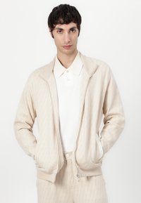 Bežna zip-up jakna z geometrijskim vzorcem, mehka pletenina, lahka zasnova in ovratnik stojač. V kombinaciji z belim teksturiranim polo majico.
