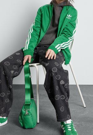 Osoba siedząca w zielonej kurtce Adidas, szarej bluzie z kapturem, czarnych spodniach z motywem uśmiechniętych buziek, zielonych trampkach, trzymająca zieloną torbę za pasek.