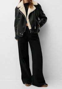 Giacca in pelle nera con colletto in shearling crema, chiusura con zip e tessuto testurizzato. Abbinata a pantaloni neri larghi e scarpe nere.