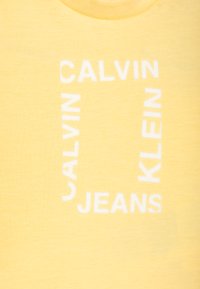 Žluté bavlněné tričko s bílým blokovým textem "CALVIN KLEIN JEANS" uspořádaným v obdélníkovém vzoru. Hladká textura.