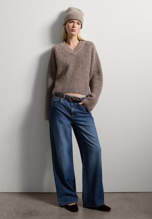 Brauner, strukturierter Strickpullover mit V-Ausschnitt und lockerem Schnitt, kombiniert mit weit geschnittenen blauen Denim-Jeans und einer passenden braunen Mütze.