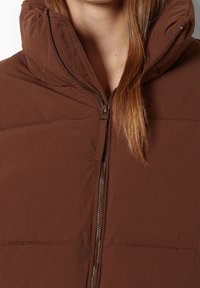 Gilet isolato marrone con colletto alto e chiusura a zip. Il materiale sembra lisciato con una leggera lucentezza, caratterizzato da dettagli di cuciture orizzontali.