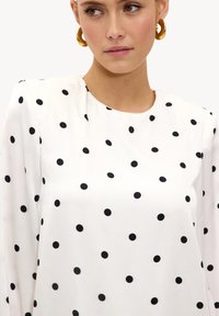 Blouse blanche présentant un motif à pois avec des points noirs. Le tissu semble lisse, et le modèle comporte un col rond ainsi que des manches longues.