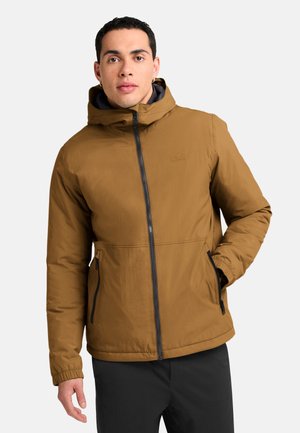 Jonge man met een bruine winterjas met capuchon, ritszakken en een zwarte broek tegen een effen achtergrond.