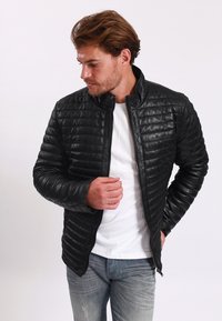 Giacca nera trapuntata in pelle sintetica, con colletto alto e zip frontale. Indossata sopra una camicia bianca con jeans azzurri.