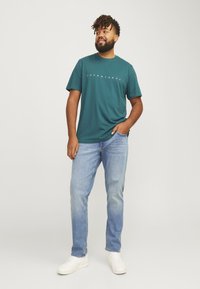 Teal cotton t-shirt with white logo text, paired with light blue jeans and white sneakers. Casual fit and short sleeves.