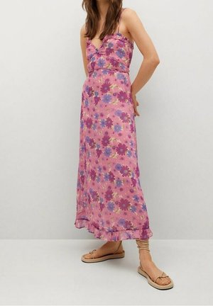 Kobieta w różowej, kwiatowej sukni maxi na cienkich ramiączkach i beżowych sandałach na sznurowadła, stojąca na tle jednolitego białego tła.