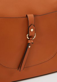 Sac en similicuir marron avec une fermeture à rabat, des surpiqûres noires et un accent en métal doré. Comprend une bandoulière décorative avec un anneau.