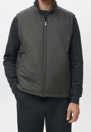 Homme portant un gilet zippé gris foncé sur un pull à manches longues foncé, les mains dans les poches du pantalon, sur un fond uni.