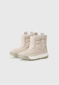 Bottes matelassées beige clair avec un dessus rembourré et une semelle texturée, dotées de poignées pour un enfilage facile. Marque visible sur le côté.