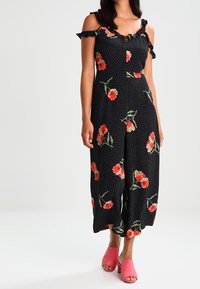 Czarny kombinezon w białe grochy, z kwiatowym nadrukiem, falbankami na ramionach i krótkimi szerokimi nogawkami. Noszony z różowymi sandałami na obcasie z odkrytymi palcami.