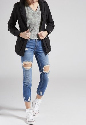 Persona con giacca nera, maglia grigia, jeans blu strappati e scarpe da ginnastica bianche, in piedi su uno sfondo semplice.