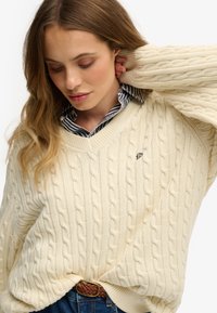 Suéter de punto trenzado en color crema con escote en V, que presenta un pequeño logo en el pecho. Lleva puesto sobre una camisa a rayas azul y blanca, combinada con pantalones de mezclilla.