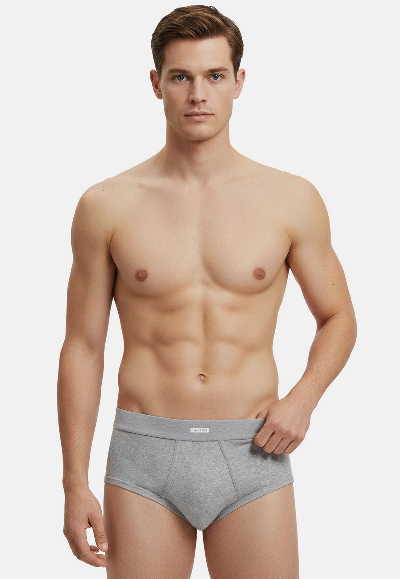 Uomo senza maglietta con slip grigi con fascia in vita con marchio, in piedi davanti a uno sfondo bianco neutro, che guarda dritto davanti a sé.