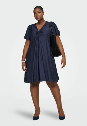 Žena v tmavomodrých krátkych šatách s viazaním vpredu, tmavomodré lodičky, perlový náramok, dlhé náušnice a veľká čierna kabelka cez rameno.