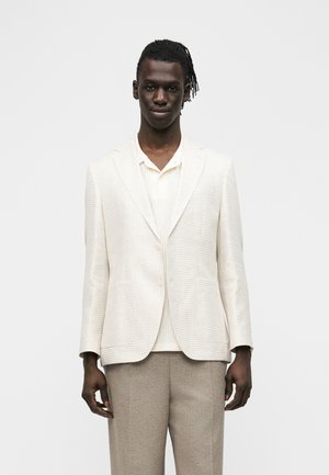 Mann trägt ein hellbeiges, strukturiertes Sakko, ein weißes Strickshirt und taupefarbene Hose, steht vor einem schlichten weißen Hintergrund.