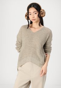 Maglione lavorato a maglia beige con scollatura a V, caratterizzato da un motivo testurizzato e polsini a costine. Abbinato a pantaloni tapered di colore chiaro.