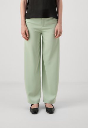 Pantaloni a gamba larga in morbido tessuto verde menta, caratterizzati da una texture liscia e dettagli di pieghe. Abbinati a scarpe nere con punta quadrata.