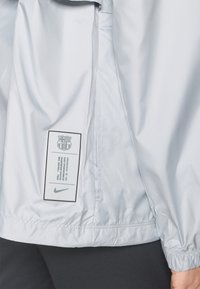 Veste coupe-vent gris clair avec des poignets élastiques, texture lisse, et une étiquette avec un logo et du texte sur la couture latérale.