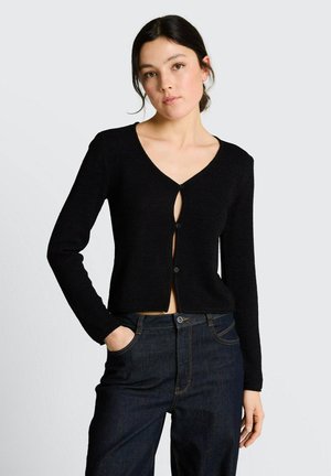 Kobieta w czarnym, długim rękawem, krótkim kardiganie zapinanym na guziki na środku oraz w ciemnoniebieskich, wysokich spodniach jeansowych, stojąca z jedną ręką w kieszeni.