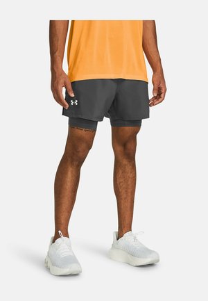 Mandlig atlet iført sorte lagdelte Under Armour løbeshorts, orange skjorte og hvide løbesko står mod en hvid baggrund.