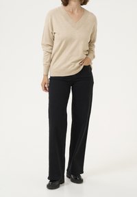 Pull beige à col en V avec manches longues, associé à un pantalon noir à jambes larges. Le mannequin porte des chaussures noires à semelles épaisses.