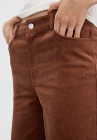 Bruna kordflängdsbyxor med vertikala ränder, med en framficka och en metallknapp. Närbild av tygtextur och passform runt midjan.