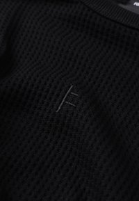 Camisola preta texturada com design canelado e logótipo subtil em contraste na parte da frente. Tecido leve, decote redondo e mangas curtas.