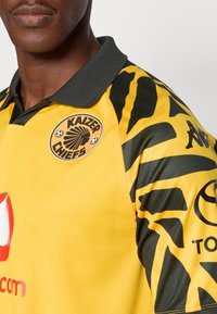 Κίτρινη φανέλα ποδοσφαίρου με μαύρο φυλετικό σχέδιο στα μανίκια, σκούρο γιακά και λογότυπα της Kaizer Chiefs και των χορηγών.