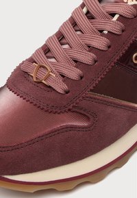 Gros plan d'une basket bordeaux en tissu structuré et cuir, lacets assortis, et un petit accessoire métallique près de la zone des orteils.